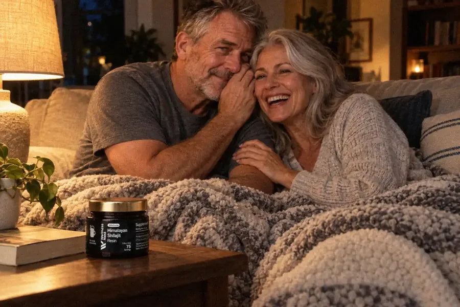 A man of about 62 whispers in his wife's ear on a sofa at night under one blanket, she laughs out loud, the ShilaSource jar on the coffee table beside a paperback