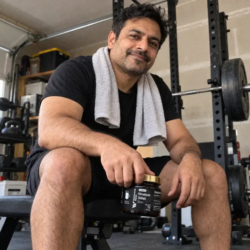 A South Asian man of about 43 with a towel round his neck sits on a gym bench in his garage, the ShilaSource jar hanging from one hand, a steady smile